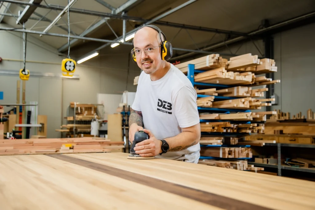 Een medewerker houtwerkplaats kijkt in de camera met een glimlach. Hij heeft een schuurmachine vast.